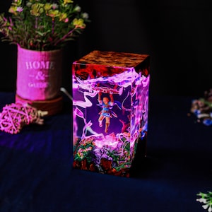 May include: A square resin lamp featuring a miniature figure suspended within. The lamp has a wooden top and a vibrant, glowing interior with shades of purple, red, and white. The base of the lamp has a landscape design.