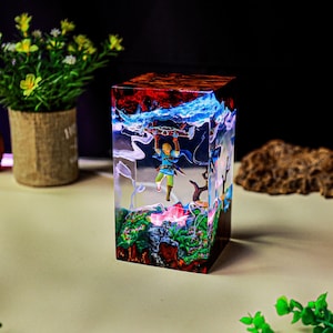 May include: A clear resin cube featuring a miniature figure of a character holding a glider. The cube has a colorful interior with red, blue, and green elements, resembling a fantasy landscape. The cube is a decorative item.