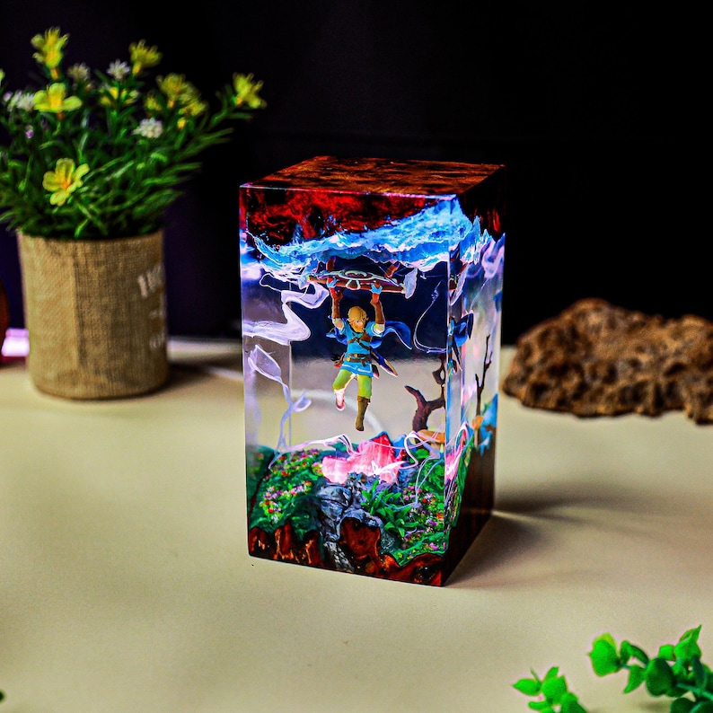 May include: A clear resin cube featuring a miniature figure of a character holding a glider. The cube has a colorful interior with red, blue, and green elements, resembling a fantasy landscape. The cube is a decorative item.