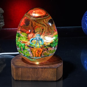 Puede incluir: Una l&aacute;mpara de vidrio en forma de huevo con una escena colorida en su interior. La escena presenta una figura de pelo azul de pie en un entorno de bosque. La l&aacute;mpara est&aacute; sobre una base de madera.