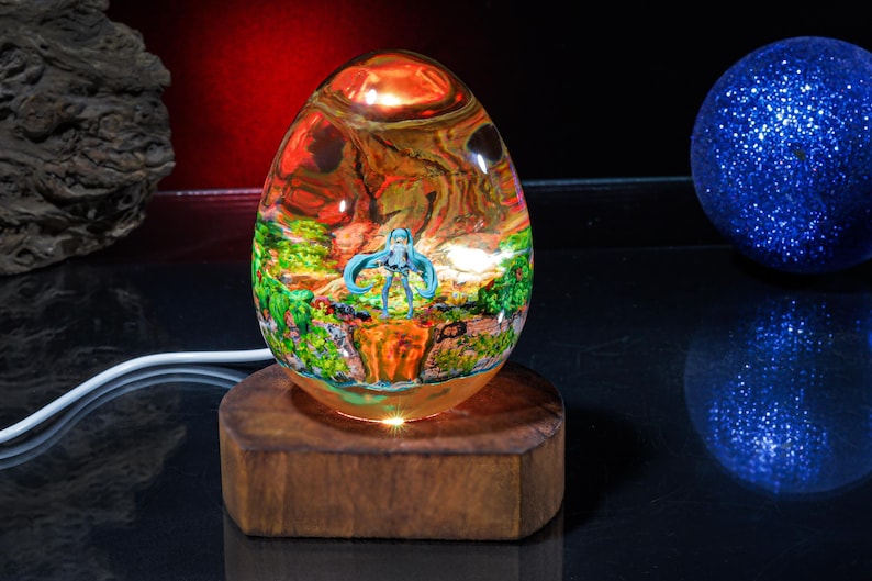 Puede incluir: Una l&aacute;mpara de vidrio en forma de huevo con una escena colorida en su interior. La escena presenta una figura de pelo azul de pie en un entorno de bosque. La l&aacute;mpara est&aacute; sobre una base de madera.