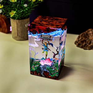 May include: A rectangular resin art piece featuring a miniature figure of a character from a video game. The top of the piece has a red and brown pattern, with blue and white accents. The figure is suspended within the clear resin, above a colorful landscape.