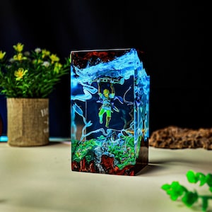 May include: A rectangular resin sculpture featuring a miniature figure holding onto a log, set within a landscape of green and red. The sculpture has blue and white accents, resembling water and clouds.