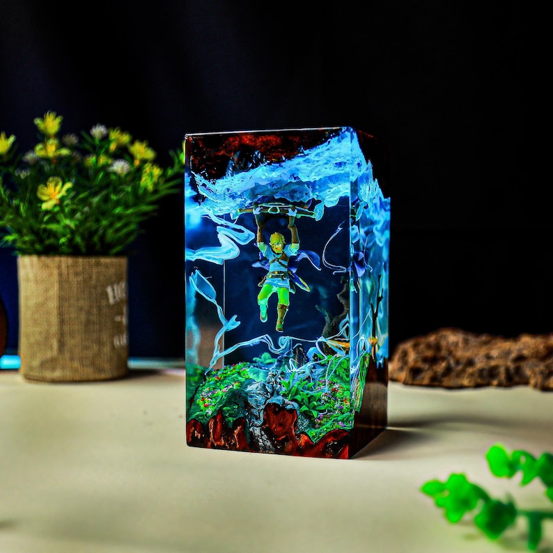 May include: A rectangular resin sculpture featuring a miniature figure holding onto a log, set within a landscape of green and red. The sculpture has blue and white accents, resembling water and clouds.