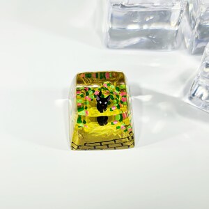 K&ouml;nnte beinhalten: Ein transparenter gelber Keycap mit einer darin gemalten schwarzen Katzenfigur und gr&uuml;nen und rosa floralen Details. Der Keycap hat ein Ziegelmuster am Boden.