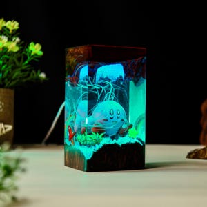 May include: A rectangular resin night light featuring a cartoon character in a glowing blue and green underwater scene. The wooden base has a dark brown color, and the light emits a soft glow.