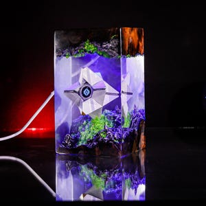 May include: A rectangular resin lamp featuring a white geometric ghost-like figure suspended inside. The base and top contain dark wood elements with bright green moss-like accents. The interior glows with a vibrant purple light.