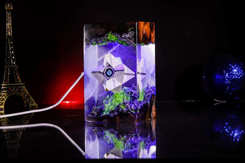 May include: A rectangular resin lamp featuring a white geometric ghost-like figure suspended inside. The base and top contain dark wood elements with bright green moss-like accents. The interior glows with a vibrant purple light.