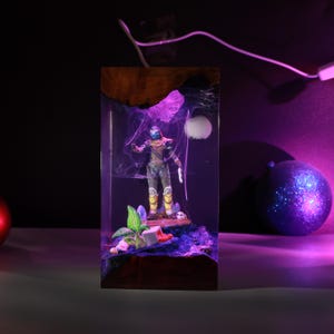 May include: A rectangular resin lamp featuring a wooden base and top, enclosing a miniature figure of a character in armor. The interior is illuminated with purple light, showcasing a small green plant, a white sphere, and a detailed landscape scene.