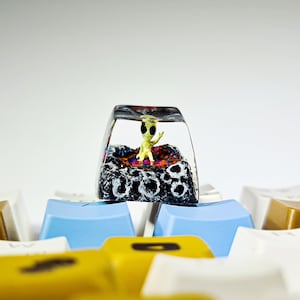 May include: A clear resin keycap with a small green alien figurine inside. The alien is sitting on a black and white textured surface. The keycap is surrounded by other keyboard keys.