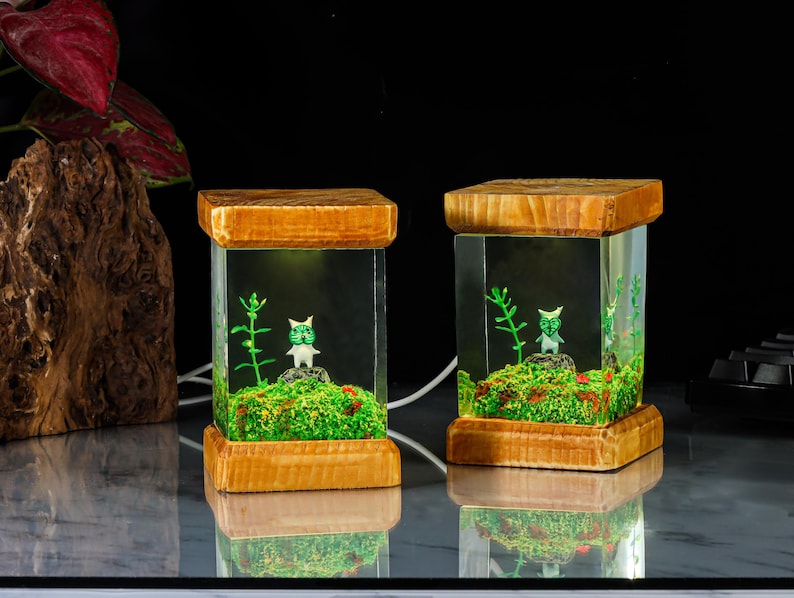 K&ouml;nnte beinhalten: Zwei gl&auml;serne Terrarien mit Holzrahmen, in denen sich eine gr&uuml;ne Pflanze und eine kleine wei&szlig;e Figur befinden. Die Terrarien sind von innen beleuchtet und strahlen ein warmes Licht aus.