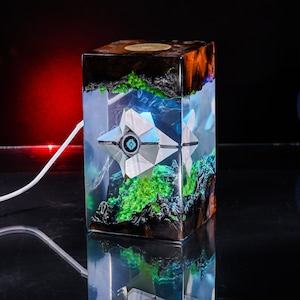 May include: A rectangular resin lamp featuring a white, geometric, star-shaped figure suspended inside. The clear resin is accented with dark wood at the top and bottom, with vibrant green moss-like textures surrounding the central white figure.