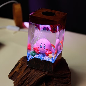May include: A unique, handmade tissue box featuring a pink Kirby character inside a clear resin cube. The cube has a wooden top and base, with a colorful rainbow and star accents. The design is illuminated with a soft, glowing light.