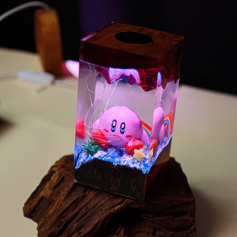 May include: A unique, handmade tissue box featuring a pink Kirby character inside a clear resin cube. The cube has a wooden top and base, with a colorful rainbow and star accents. The design is illuminated with a soft, glowing light.