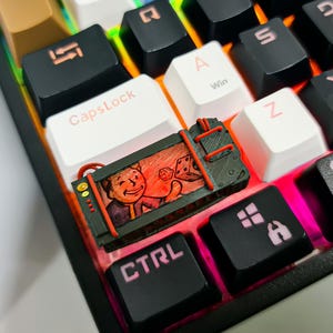 May include: A black and red keyboard keycap with a cartoon image of a person holding a dice. The keycap is on a black and white keyboard with rainbow lighting.