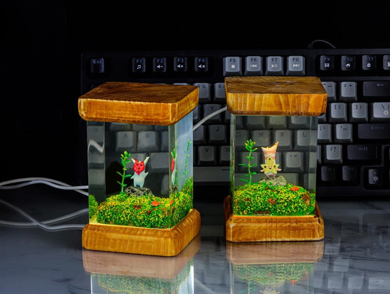 K&ouml;nnte beinhalten: Zwei gl&auml;serne Terrarien mit Holzrahmen, in denen sich eine kleine gr&uuml;ne Pflanze und eine kleine Figur befinden. Die Terrarien sind von innen beleuchtet und stehen auf einer schwarzen Tastatur.