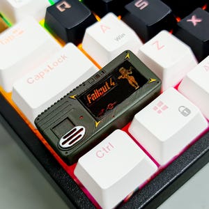 Op de afbeelding: Een olijfgroene keycap met gele details, met het Fallout 4-logo en een karakter. De keycap staat tussen witte en zwarte toetsenbordtoetsen. Een aangepast accessoire voor gamers.