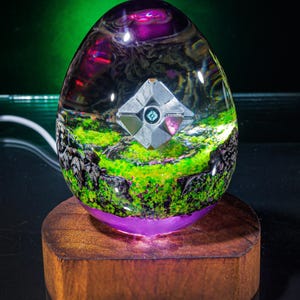 May include: An egg-shaped resin sculpture with a silver geometric object inside, set on a wooden base. The sculpture features a landscape with green and purple hues, and a glowing purple base. The top of the egg is a dark purple colour.