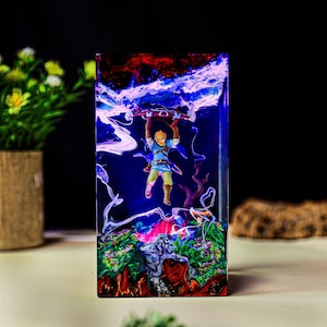 May include: A rectangular resin art piece featuring a miniature figure of a character from The Legend of Zelda. The figure is suspended in a vibrant scene with blue, red, and white colors, depicting a stormy sky and a landscape below. The piece is approximately 6 inches tall.