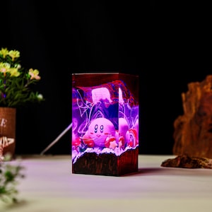 May include: A rectangular wooden lamp featuring a pink cartoon character with a smiling face. The lamp has a vibrant purple and red interior with a textured appearance. The lamp is on a white surface.