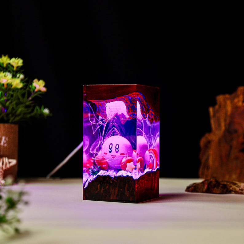 May include: A rectangular wooden lamp featuring a pink cartoon character with a smiling face. The lamp has a vibrant purple and red interior with a textured appearance. The lamp is on a white surface.