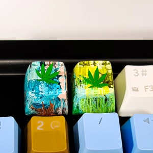May include: Two custom keyboard keycaps with a green marijuana leaf design. The keycaps are made of clear resin with a swirling blue, green, and brown pattern. The keycaps are on a keyboard with yellow, blue, and white keys.