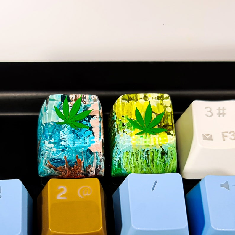 May include: Two custom keyboard keycaps with a green marijuana leaf design. The keycaps are made of clear resin with a swirling blue, green, and brown pattern. The keycaps are on a keyboard with yellow, blue, and white keys.