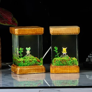 K&ouml;nnte beinhalten: Zwei Holztischlampen mit klaren Glasschirmen. Jede Lampe hat eine kleine gr&uuml;ne oder gelbe Figur in einem Miniaturterrarium mit Moos und Gr&uuml;n.