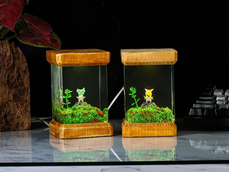 K&ouml;nnte beinhalten: Zwei Holztischlampen mit klaren Glasschirmen. Jede Lampe hat eine kleine gr&uuml;ne oder gelbe Figur in einem Miniaturterrarium mit Moos und Gr&uuml;n.
