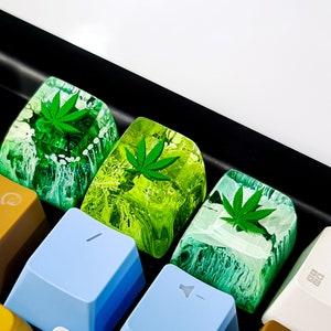 May include: Four green and white keycaps with a marijuana leaf design. The keycaps are on a black keyboard.