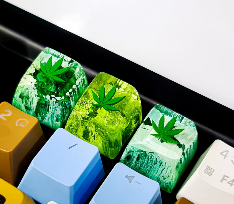 May include: Four green and white keycaps with a marijuana leaf design. The keycaps are on a black keyboard.