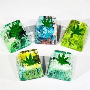 May include: Five resin keycaps with abstract designs and green marijuana leaf accents. The keycaps are in various colors, including blue, green, yellow, and pink.