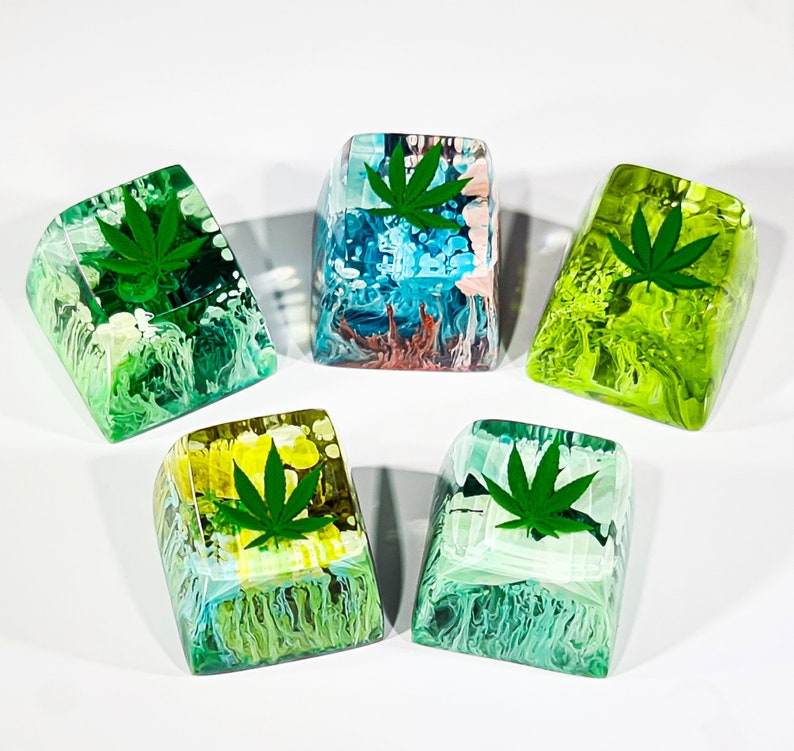 May include: Five resin keycaps with abstract designs and green marijuana leaf accents. The keycaps are in various colors, including blue, green, yellow, and pink.