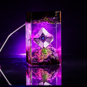 May include: A rectangular resin lamp featuring a white geometric Destiny Ghost shell suspended inside. The clear resin block contains dark textured elements at the top and bottom, illuminated by a vibrant purple LED light source.