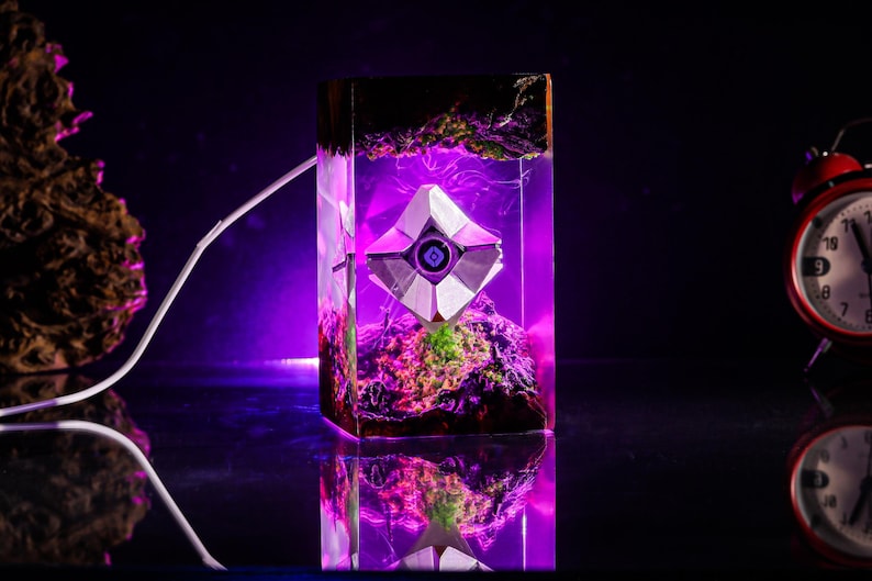 May include: A rectangular resin lamp featuring a white geometric Destiny Ghost shell suspended inside. The clear resin block contains dark textured elements at the top and bottom, illuminated by a vibrant purple LED light source.