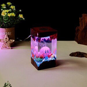 May include: A decorative, illuminated cube featuring a pink cartoon character inside a clear resin block. The cube has a dark wood-like top and base, with colorful accents and a soft, glowing interior. The character is surrounded by miniature aquatic elements.