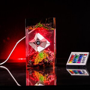May include: A rectangular resin lamp containing a geometric white robotic figure suspended within a red and green landscape. The lamp features a white power cord and is accompanied by a small remote control with multiple colored buttons.