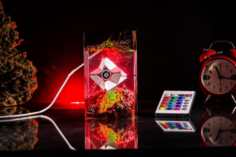 May include: A rectangular resin lamp containing a geometric white robotic figure suspended within a red and green landscape. The lamp features a white power cord and is accompanied by a small remote control with multiple colored buttons.