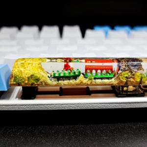 May include: A custom artisan keycap featuring a miniature landscape scene. The keycap is clear with a detailed diorama of a bridge, trees, and figures. The keycap is set on a white and blue mechanical keyboard.