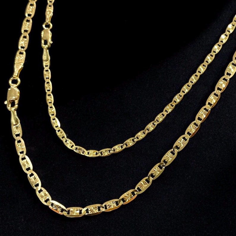 May include: Two gold chains with a mariner link pattern. The chains are layered on top of each other and have a textured surface.
