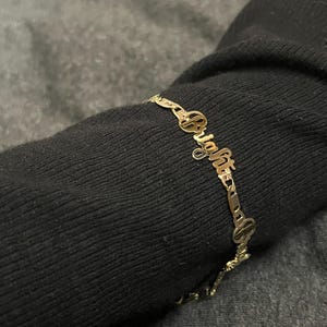 May include: A gold-colored bracelet with the name "Bright" spelled out in cursive script. The bracelet is made of delicate links and is worn on a dark sleeve. The word "Bright" is the focal point of the jewelry.