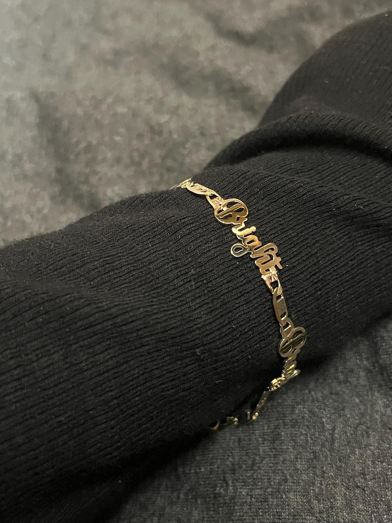 May include: A gold-colored bracelet with the name "Bright" spelled out in cursive script. The bracelet is made of delicate links and is worn on a dark sleeve. The word "Bright" is the focal point of the jewelry.