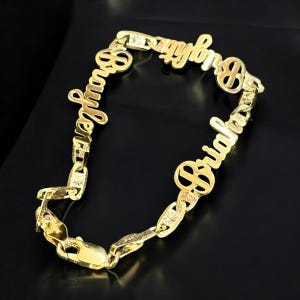 May include: A gold-colored name bracelet with the names "Braylen" and "Briah" in a cursive font. The bracelet has a chain link design with a clasp closure. The background is black.