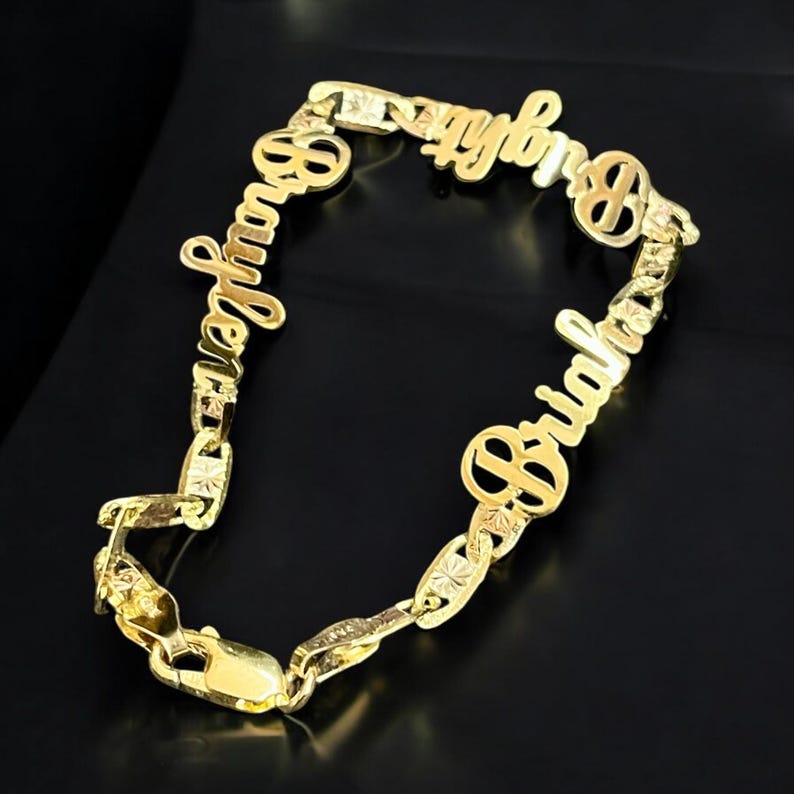 May include: A gold-colored name bracelet with the names "Braylen" and "Briah" in a cursive font. The bracelet has a chain link design with a clasp closure. The background is black.