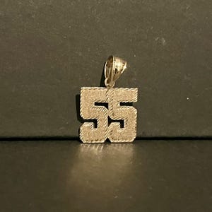 May include: Gold-toned pendant in the shape of the number 55. The pendant has a textured surface and a loop at the top for a chain. The background is a dark, neutral color.