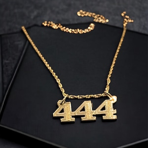 Könnte beinhalten: Eine goldene Kettenkette mit einem Anhänger mit der Zahl 444. Die Kette wird auf einem schwarzen Hintergrund präsentiert.