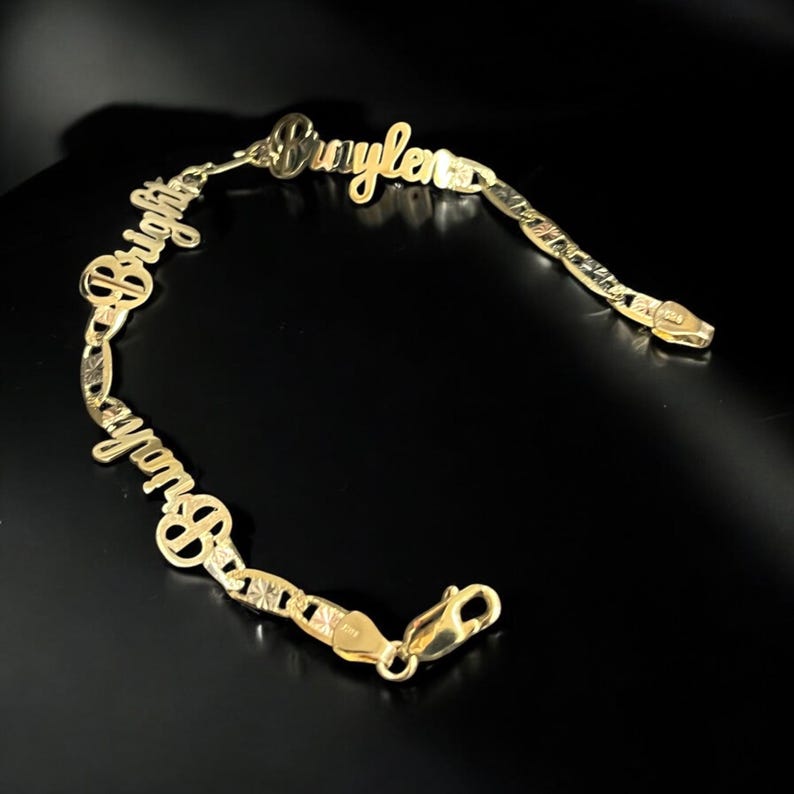 May include: A gold-colored bracelet with the names "Brightyn" and "Braylen" in a cursive font. The bracelet has a lobster clasp closure and a chain-link design with starburst accents. The background is black.