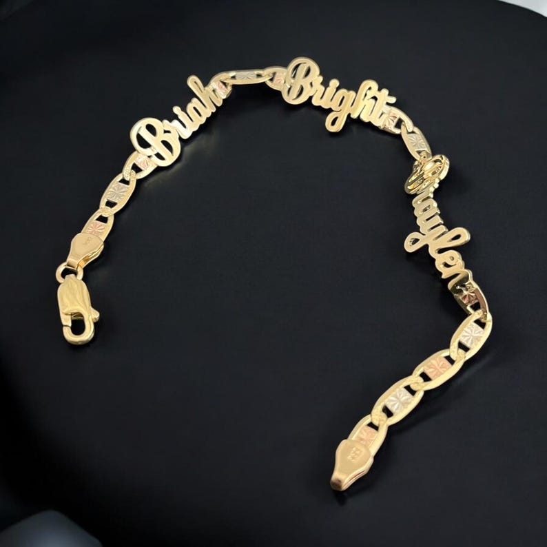 May include: A gold-colored bracelet with the names "Briaha", "Bright", and "Jaylen" spelled out in cursive script. The bracelet features a chain link design with a lobster claw clasp. The bracelet is displayed on a black surface.