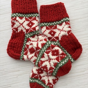 May include: Two red and white knitted Christmas stockings with a traditional Nordic pattern. The stockings have a red cuff, a white and red patterned body, and a green border. The stockings are made of a thick, textured yarn.