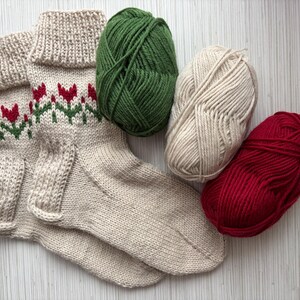 May include: A pair of beige knitted socks with a red and green floral pattern. The socks are lying on a white surface with three balls of yarn: green, beige, and red.